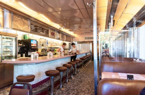 A retro diner interior with a long counter, bar stools, and two staff members preparing food.
