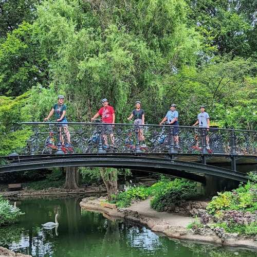 Five people on rollerblades pose on a bridge over a pond, surrounded by lush greenery and trees.