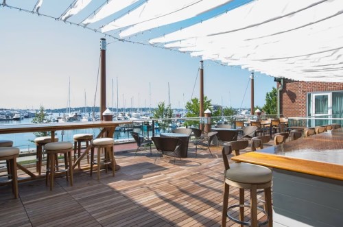 A waterfront patio with wooden flooring, bar stools, and a view of boats docked in the marina under a sunny sky.