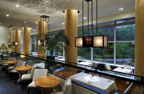 Elegant restaurant interior with wooden tables, soft lighting, and large windows overlooking greenery.