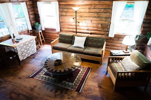 Cozy log cabin interior with wooden walls, a sofa, coffee table, and a dining area, illuminated by natural light.