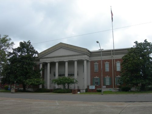 A large brick building with columns, featuring a flagpole and surrounded by trees, under a cloudy sky.