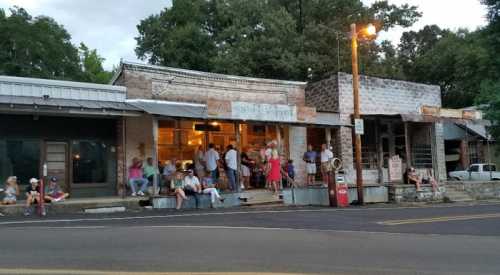 A group of people gathers outside a rustic storefront in a small town, with trees and buildings in the background.