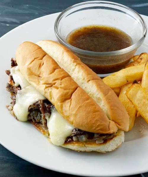 A plate with a cheesy beef sandwich, served with fries and a small bowl of dipping sauce.