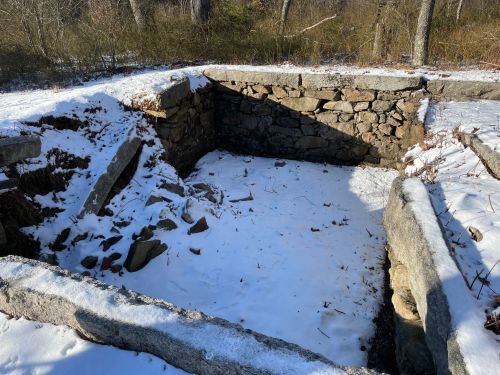 An empty stone foundation surrounded by snow and trees, with remnants of walls partially visible.