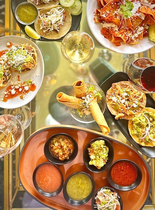 A vibrant spread of tacos, enchiladas, and various salsas, served with drinks on a glass table.