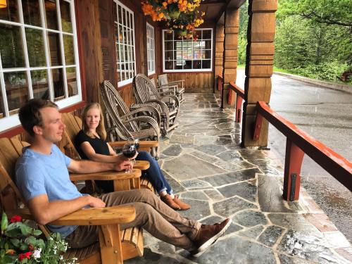 A man and woman sit on a porch in wooden chairs, enjoying drinks while it rains outside. Flowers hang above them.