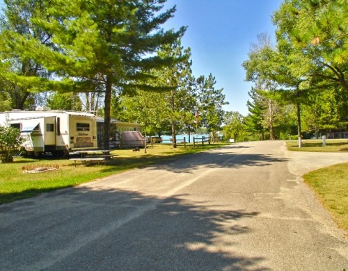 A peaceful campsite with RVs parked along a tree-lined road, leading to a lake in the background.