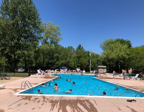 A sunny day at a public pool with children swimming and people relaxing on lounge chairs around the water.
