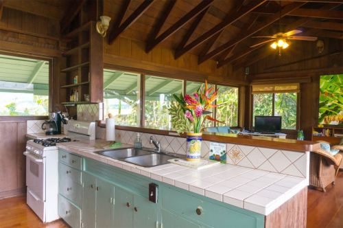 Bright kitchen with wooden beams, tiled countertops, and tropical plants visible through large windows. Cozy and inviting atmosphere.