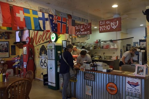 A cozy diner with flags, a welcoming sign, and staff preparing food behind the counter.