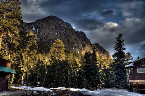 A scenic view of mountains surrounded by tall trees, with a hint of snow on the ground and dramatic clouds overhead.
