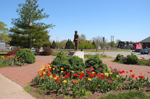 A bronze statue stands in a park surrounded by colorful flower beds and greenery under a clear blue sky.