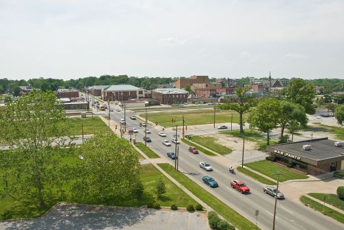 Aerial view of a small town with green spaces, buildings, and cars on the road under a clear sky.