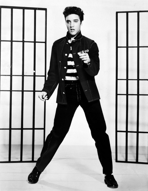 Elvis Presley poses in a striped shirt and jacket, standing between two prison bars in a black-and-white photo.