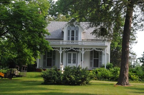 A charming white Victorian house with a porch, surrounded by lush greenery and trees.