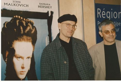 Two men stand beside a movie poster featuring a woman with a dramatic hairstyle, with names of actors above.