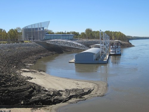 A modern building by a river, with a boat docked nearby and a clear blue sky overhead.