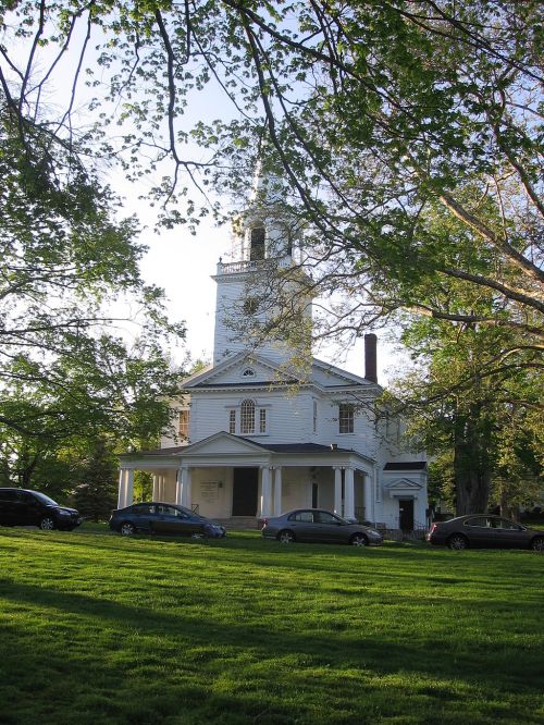 A white church with a tall steeple, surrounded by green trees and parked cars on a sunny day.