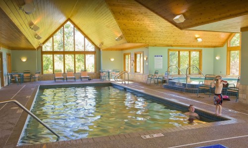 Indoor swimming pool with large windows, wooden ceiling, and two hot tubs. Two people are enjoying the water.