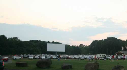 A drive-in movie theater with a large blank screen and cars parked in a grassy area under a cloudy sky.