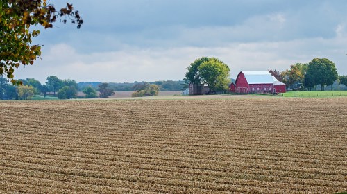 A red barn stands in a rural landscape with plowed fields and trees under a cloudy sky.