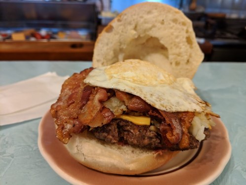 A close-up of a burger with a beef patty, melted cheese, crispy bacon, and a fried egg on a soft bun.