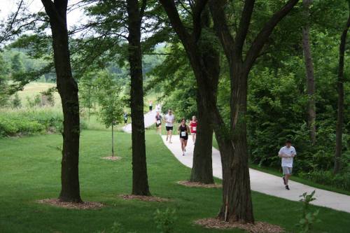 A group of runners jogs along a paved path surrounded by trees and greenery.