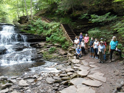 A group of hikers poses near a waterfall in a lush, green forest with stone steps in the background.