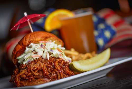 A pulled pork sandwich topped with coleslaw, served with a pickle and fries, alongside a drink with an orange slice.
