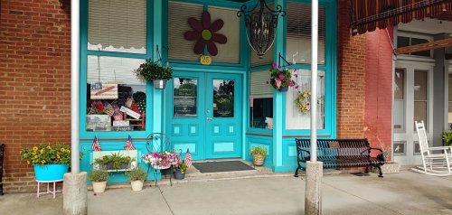 Bright turquoise storefront with flower decorations, potted plants, and American flags, featuring a welcoming entrance.