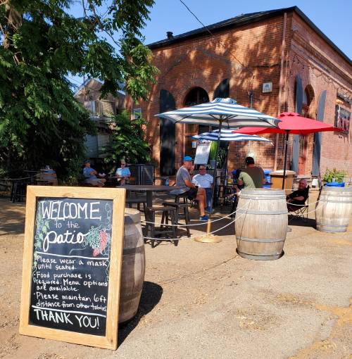 Outdoor patio with tables, umbrellas, and a welcome sign reminding guests to wear masks and maintain distance.