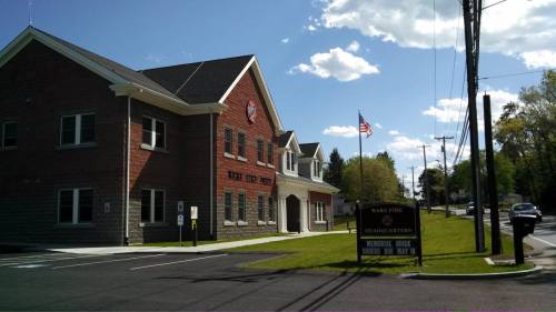 A brick fire station with a flag, surrounded by trees and a clear blue sky, located on a quiet street.