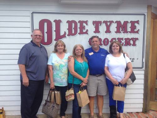 A group of five people stands in front of a sign for "Olde Tyme Grocery," smiling and holding small bags.