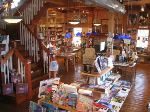 A cozy bookstore interior with wooden floors, shelves filled with books, and a staircase leading to an upper level.