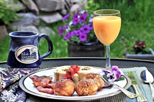 A breakfast plate with pastries, bacon, and fruit, accompanied by orange juice and coffee, set in a garden.