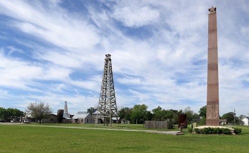 A grassy area featuring oil rigs and a tall monument under a blue sky with scattered clouds.