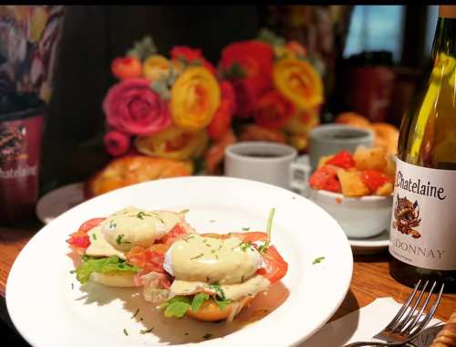 A plate of eggs Benedict with tomatoes and greens, accompanied by coffee, fruit, and a bottle of wine, with flowers in the background.