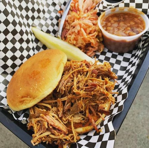 A tray with a pulled pork sandwich, coleslaw, baked beans, and a pickle, all served on a checkered paper.