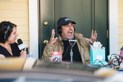A man in a cap animatedly speaks into a microphone while a woman laughs beside him, both wearing headphones.
