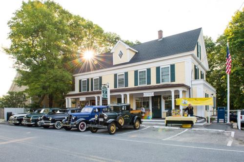 A vintage car show in front of a yellow historic building, with trees and sunlight in the background.