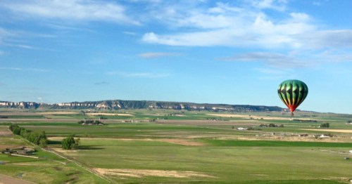 A colorful hot air balloon floats over a vast green landscape with distant hills under a clear blue sky.
