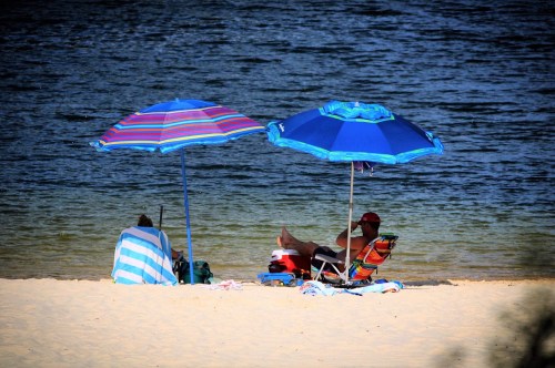 Two beach umbrellas shade people sitting on the sand by a calm water body, enjoying a sunny day.