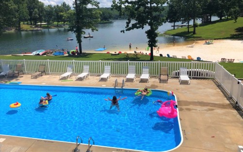 A sunny pool scene with people swimming and relaxing, surrounded by green trees and a lake in the background.