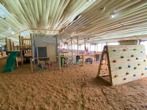 Indoor play area with climbing structures, slides, and colorful toys on a sandy floor. Brightly lit and spacious.