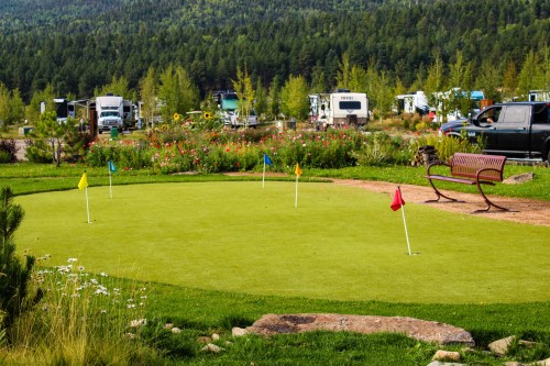 A small putting green with flags, surrounded by flowers and a bench, with RVs parked in the background.