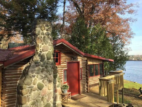 A cozy log cabin with a stone chimney, surrounded by trees and overlooking a serene lake.