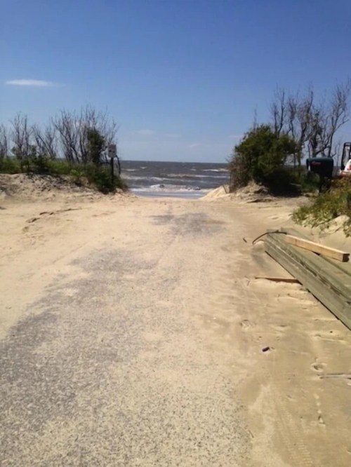 A sandy path leads to a beach with waves, framed by sparse vegetation and a clear blue sky.