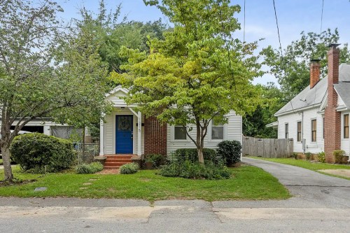 A charming single-story house with a blue door, surrounded by greenery and trees, on a quiet residential street.