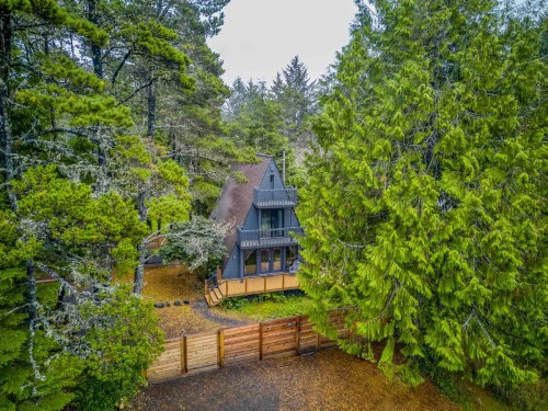 Aerial view of a cozy A-frame house surrounded by tall trees and a gravel path leading to a wooden fence.